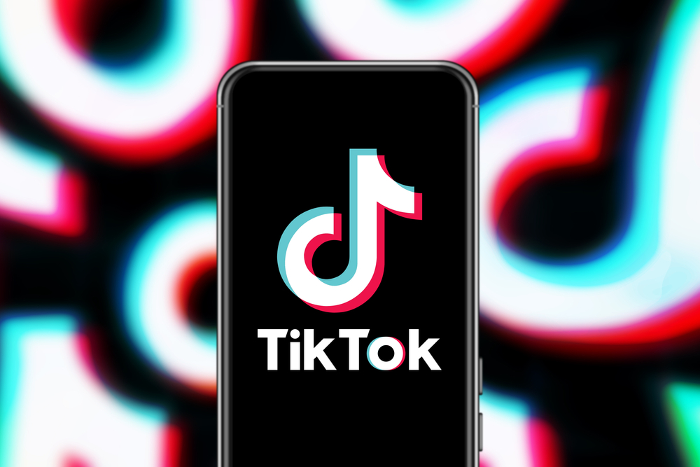 An image of an iphone with the TikTok logo enlarged on the main screen. The background of the image is the Tiktok logo but out of focus.