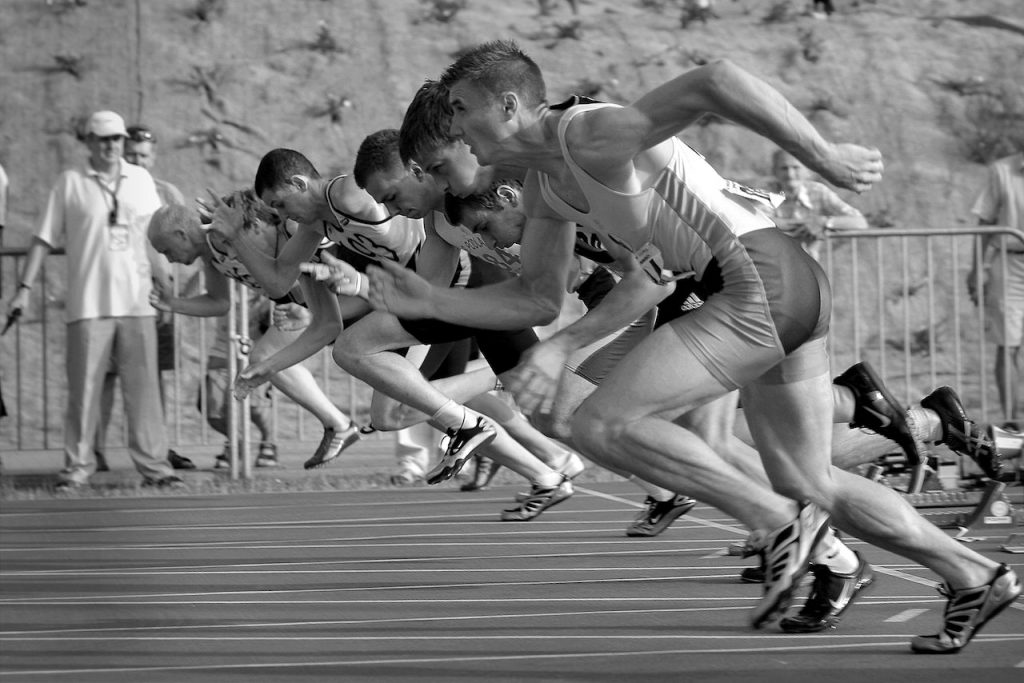 A black and white image of 7 men racing on a marathon track. They look to be in full sprint.