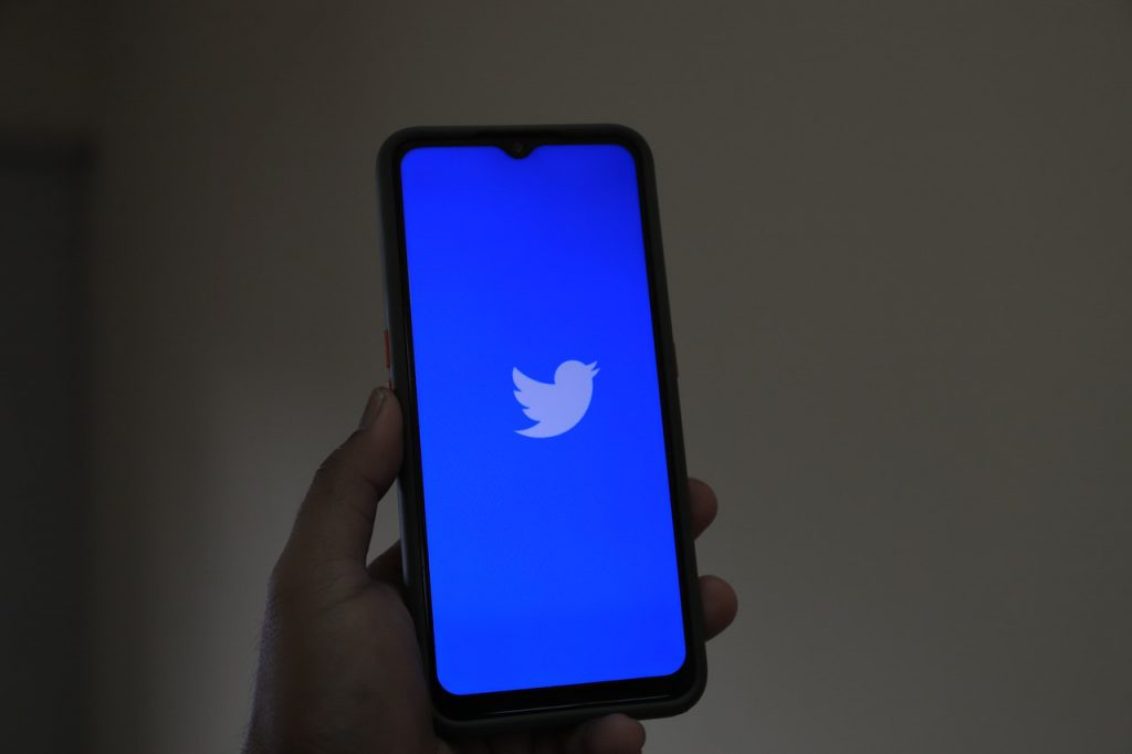 An image of an iphone with the blue twitter logo on the screen. Someone is holding the iphone to display it to the camera.