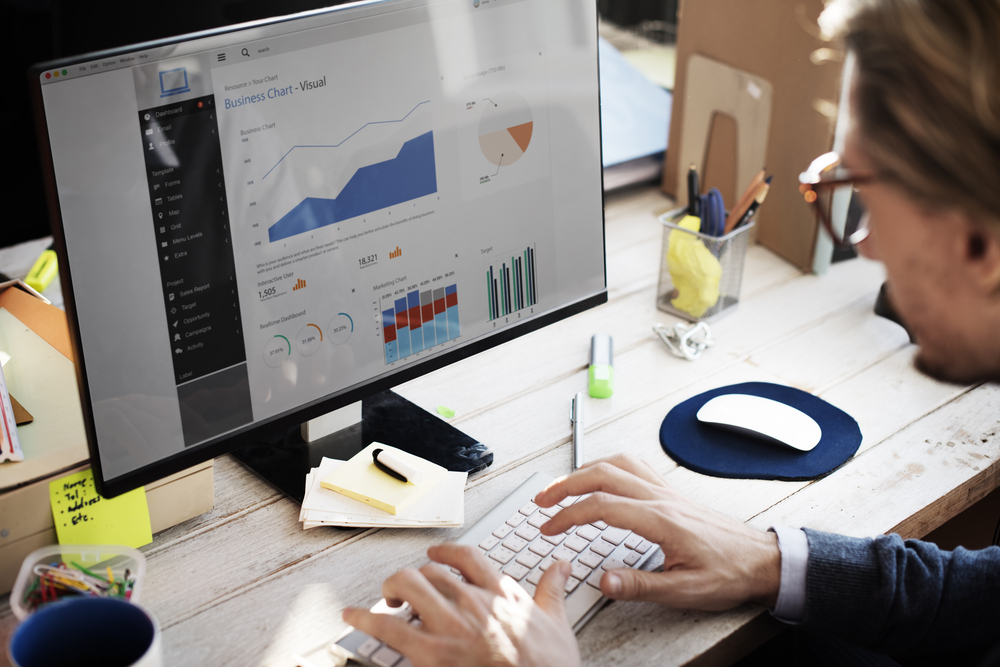 A moustached man looking at business chart analytics on a monitor, typing on a keyboard.